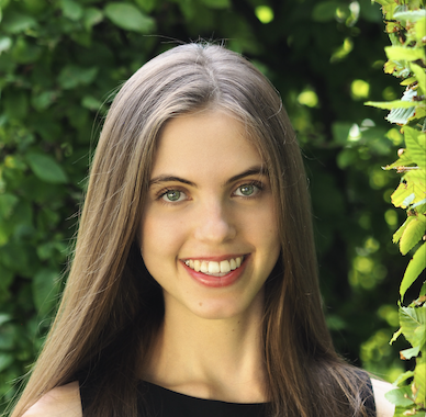 Miriam Brody with brown hair and blue/green eyes, smiling in front of a green leafy bush.